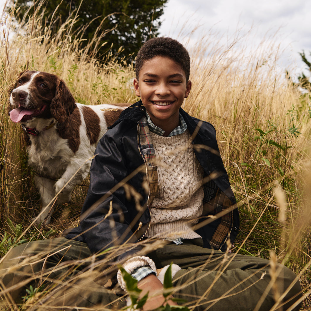 Child wearing a J.Crew sweater, button-down shirt, overcoat, and army green pants sitting in a field with a dog